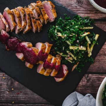 A black slate board with slices of seared duck breast, some topped with a berry sauce, beside a kale and apple salad. A small bowl of crushed red pepper flakes, a bowl of greens, and a whole red apple are arranged around the board on a wooden surface. Bookmark this setup for your dinner recipes to curb takeout addiction.