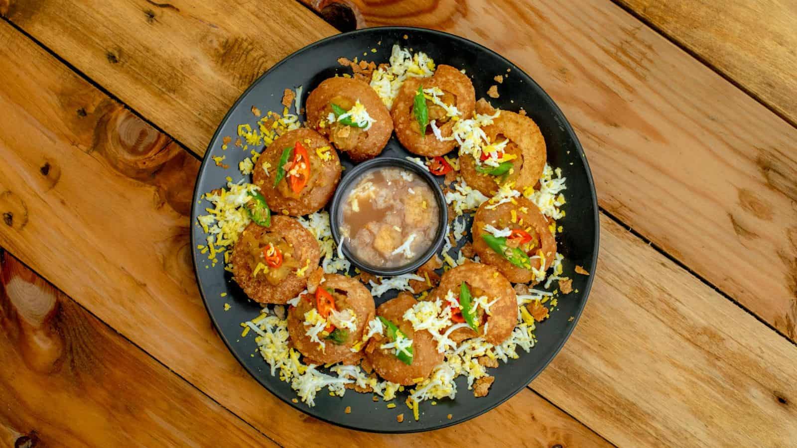 A black plate holds a ring of breaded and fried round food items, each crowned with white cheese and a sprinkle of red and green garnish, reminiscent of iconic street foods. In the center is a dipping sauce. The plate rests on a wooden surface.