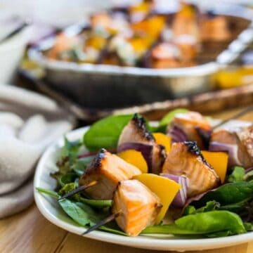 A plate of grilled salmon and vegetable skewers is placed on a bed of mixed greens. The skewers contain pieces of salmon, yellow bell pepper, and red onion. In the background, there is a tray with additional skewers and a white bowl.