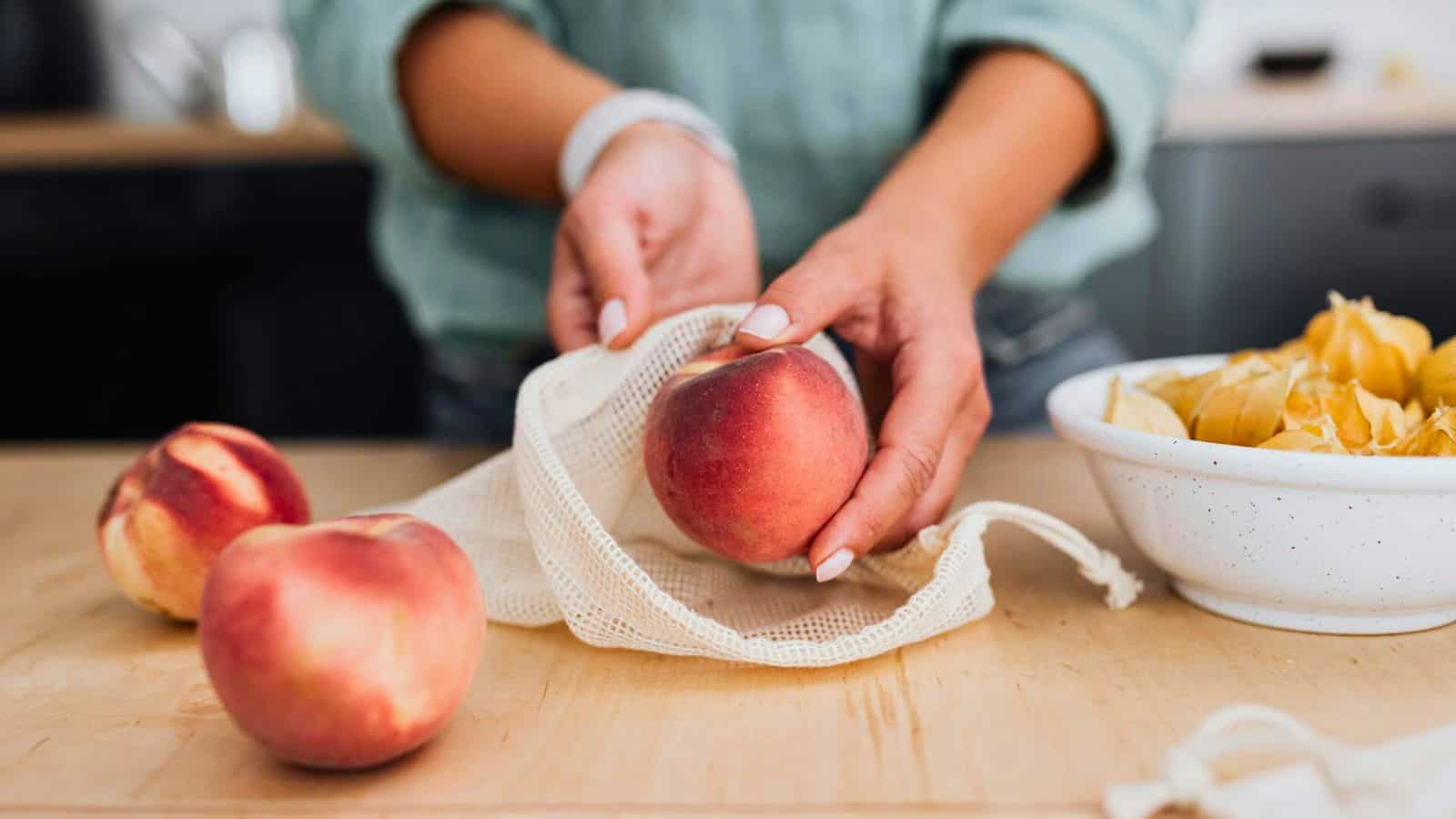 Two hands place a peach into a mesh bag on a wooden surface. Two loose peaches and a white bowl with yellow fruit husks are also on the surface.