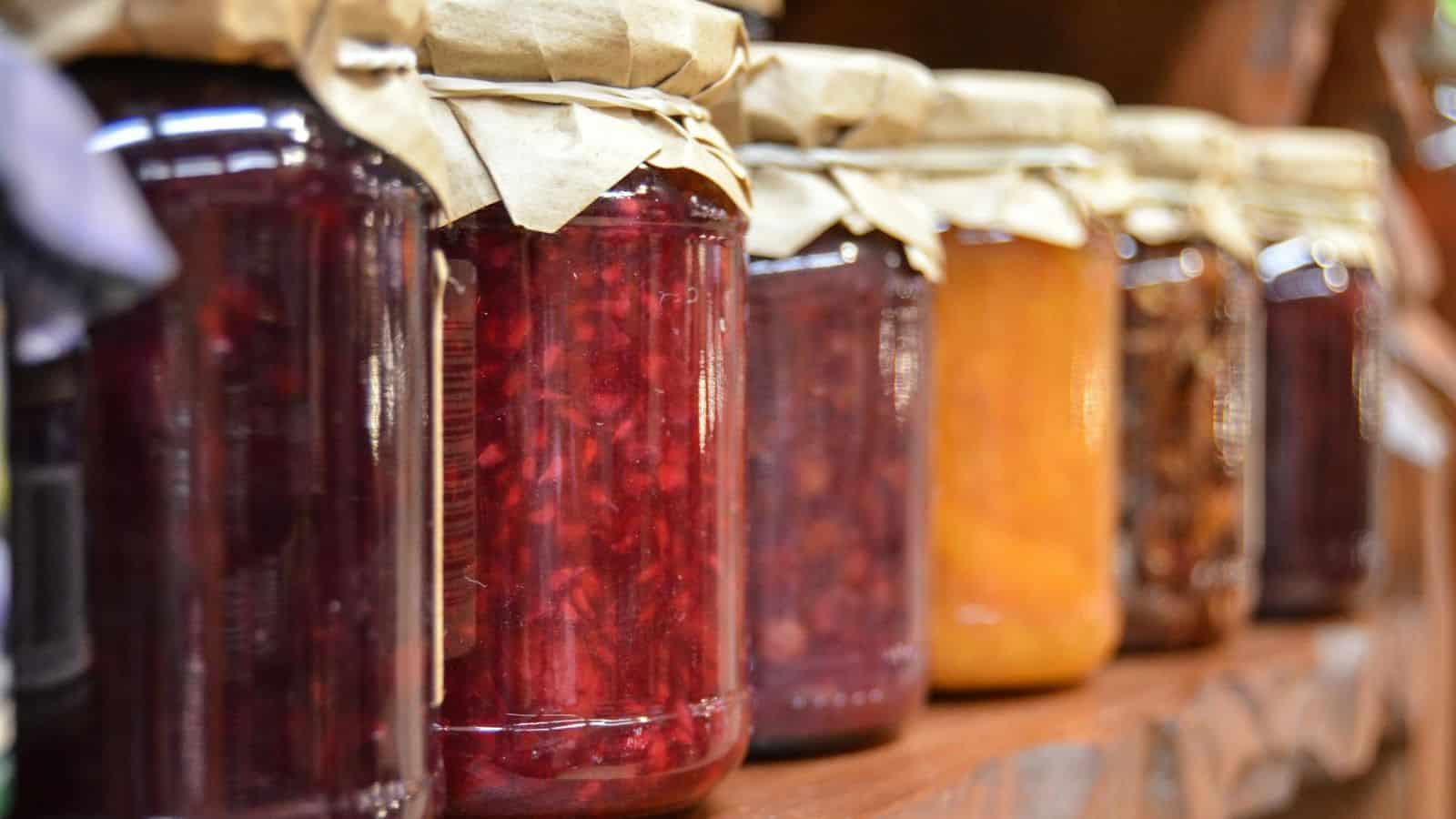 A row of glass jars filled with various homemade preserves is lined up on a wooden shelf. The jars are sealed with brown paper covers and tied with string. The preserves appear to be fruit spreads or jams in shades of red, orange, and brown.