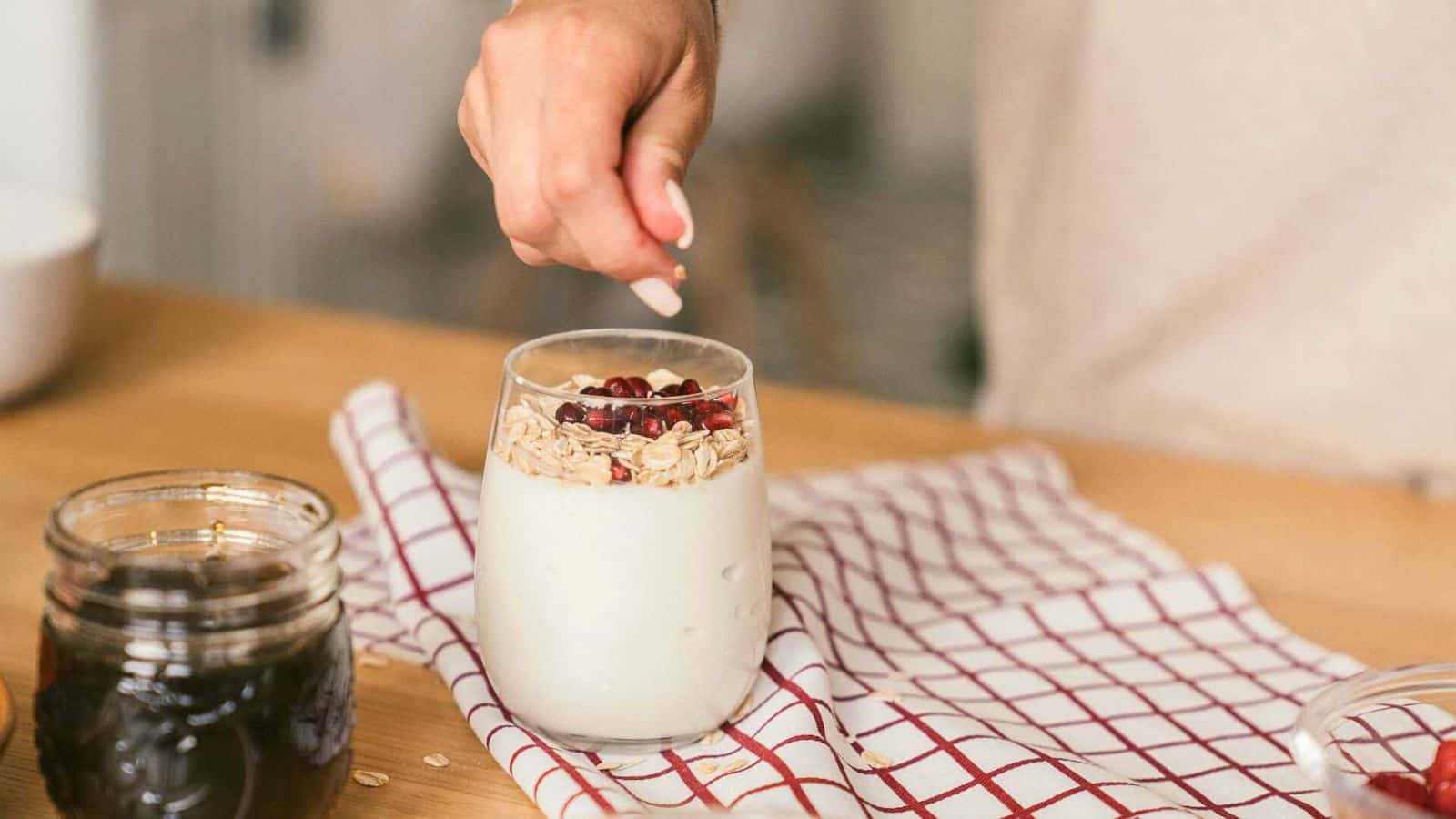 A person's hand is seen sprinkling oats or granola along with pomegranate seeds into a glass of yogurt on a wooden table. A brown mason jar filled with a dark liquid and a red-and-white checkered cloth are also visible on the table.
