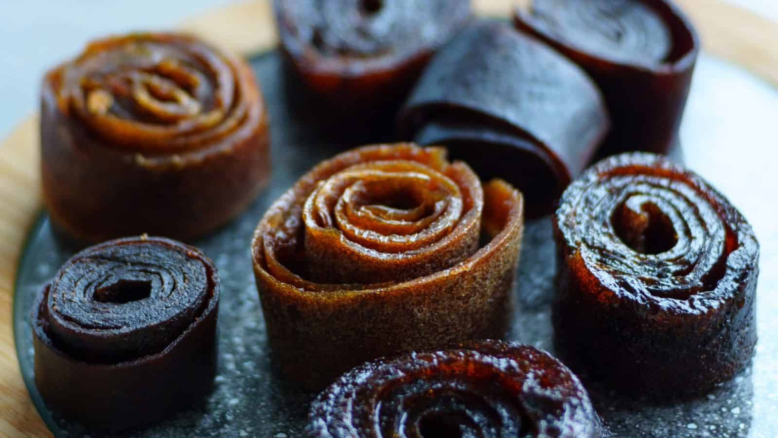 A close-up image of eight rolled fruit leather pieces in various shades of brown and dark red, arranged on a dark tray. The fruit leathers have a shiny, slightly sticky surface and are positioned in a loosely circular pattern.