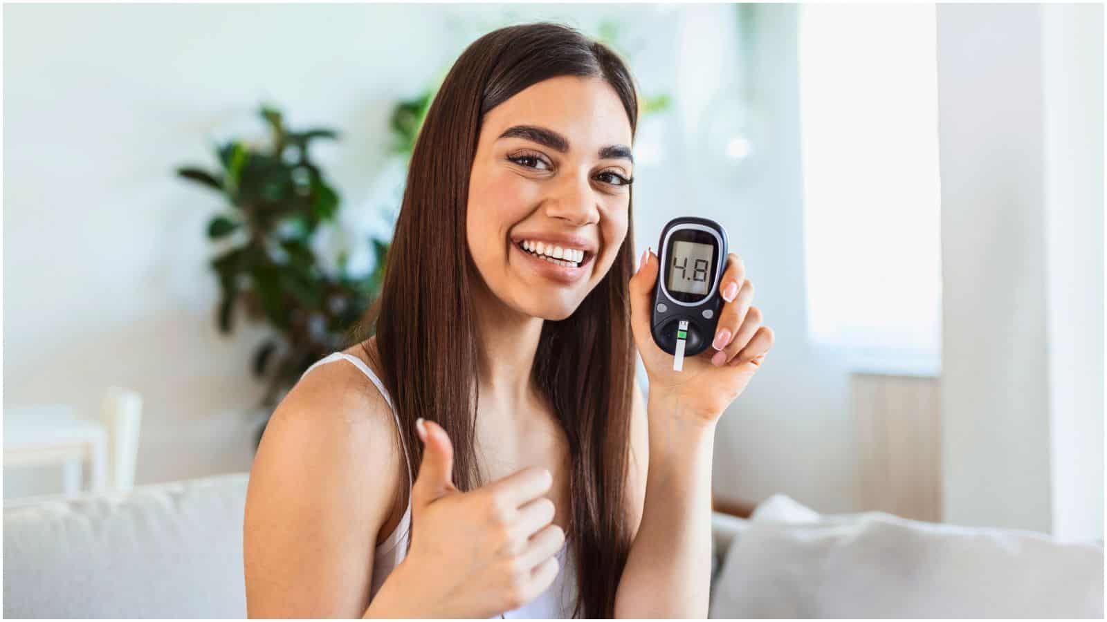 A woman with long dark hair smiles and shows a thumbs-up while holding a glucose meter displaying a reading of 4.8. She is sitting on a couch with a green plant in the background.