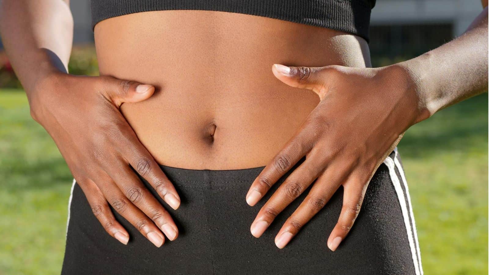 A person wearing a black crop top and black athletic pants is holding their hands on their exposed abdomen, with fingers spread out and thumbs facing inward. The background shows greenery out of focus.