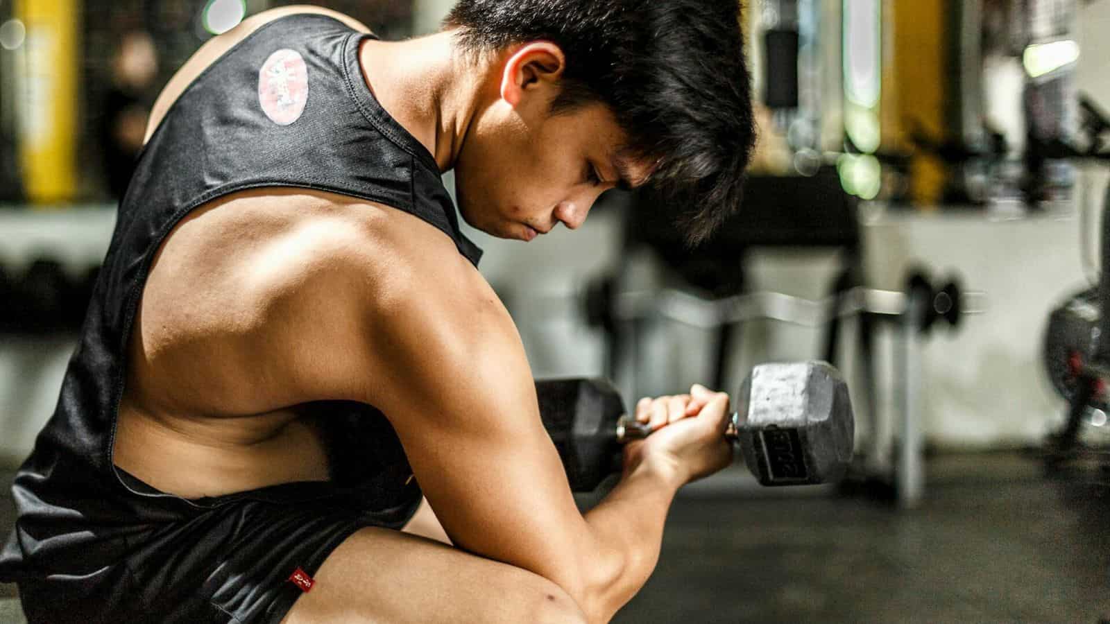 A person wearing a sleeveless shirt and shorts is sitting on a bench in a gym, intensely focusing on curling a dumbbell with their right arm. Gym equipment and weights are visible in the background.