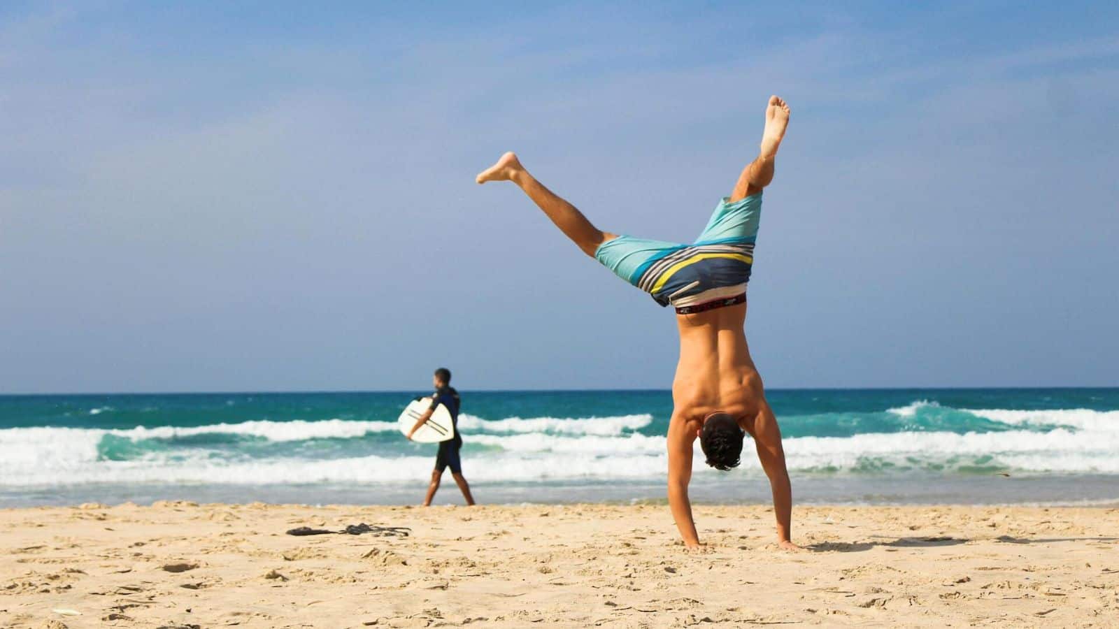 A person in swim trunks performs a handstand on a sandy beach. In the background, another person holding a surfboard walks toward the ocean, which has small waves crashing under a blue, slightly cloudy sky. 