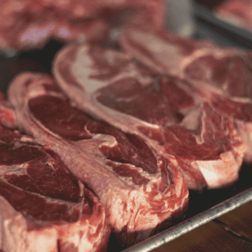 Close-up image of several raw, marbled ribeye steaks on a metal tray, ready to be prepared for cooking. The surrounding area also contains other pieces of raw meat, indicating a preparation setting. The lighting reveals the texture and fat marbling of the steaks.