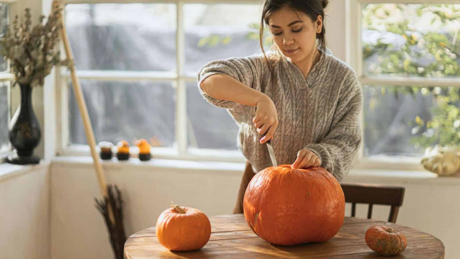 A woman in a gray sweater is carving a large pumpkin on a wooden table inside a bright room with large windows. Smaller pumpkins are also on the table, along with various items in the background, including a broom and dried plants.