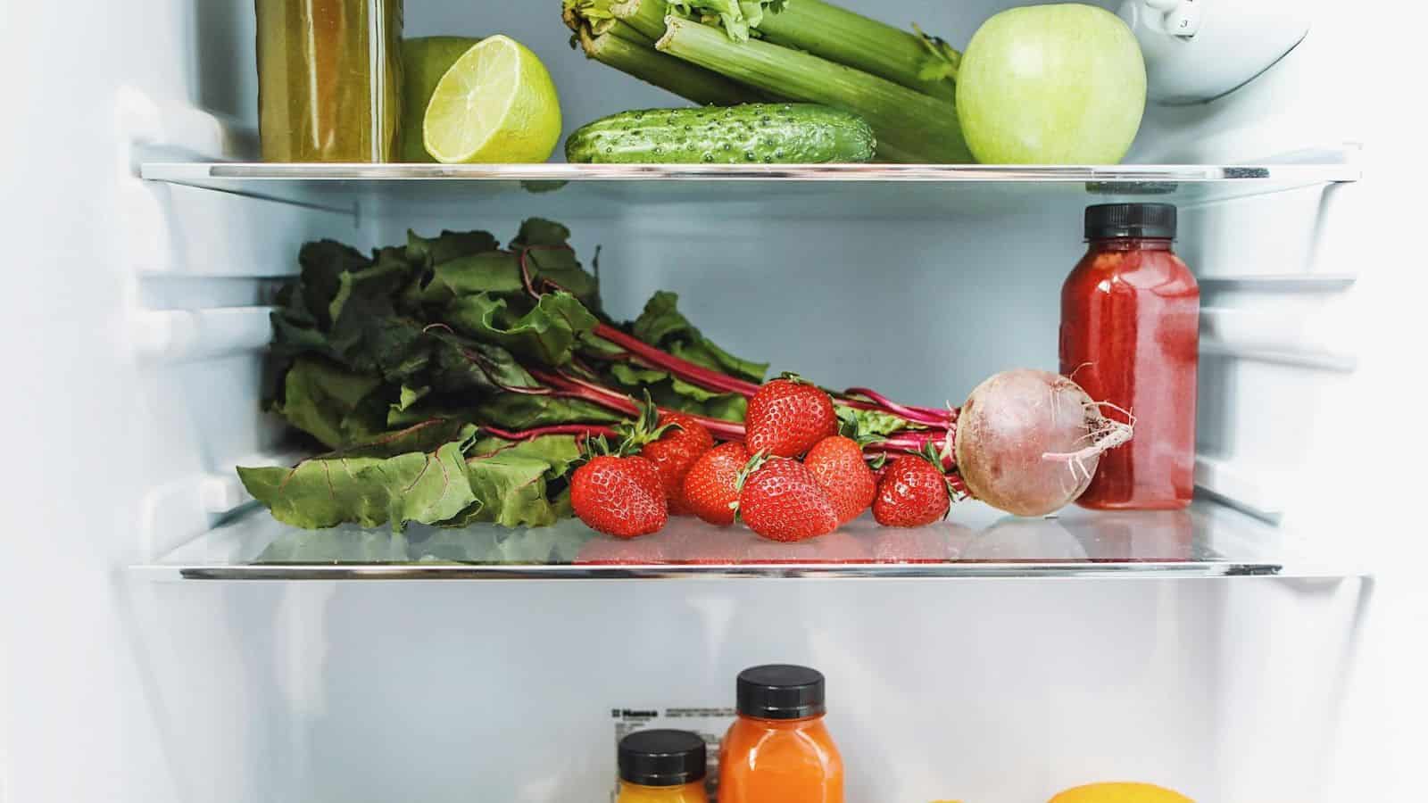 A refrigerator with neatly arranged food items including a green apple, celery, a cucumber, a lime, leafy greens, strawberries, a beetroot, a red bottle and two orange bottles alongside a few citrus fruits on the bottom shelf.