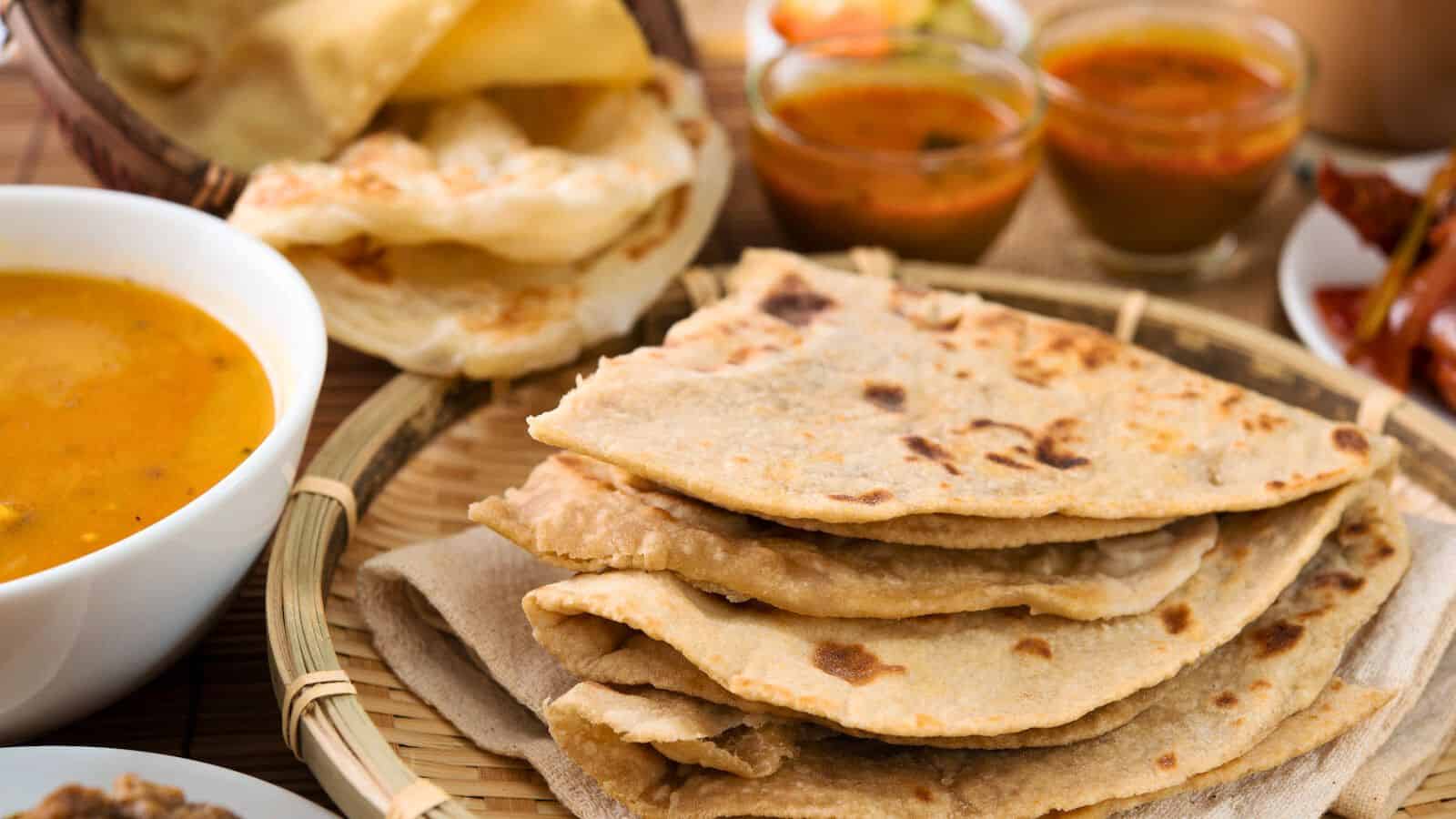 A plate of folded, golden-brown flatbreads is in the foreground. Surrounding the plate are various dishes, including bowls of curry and other side dishes. The background shows additional pieces of flatbread and a small bowl of colorful vegetables.