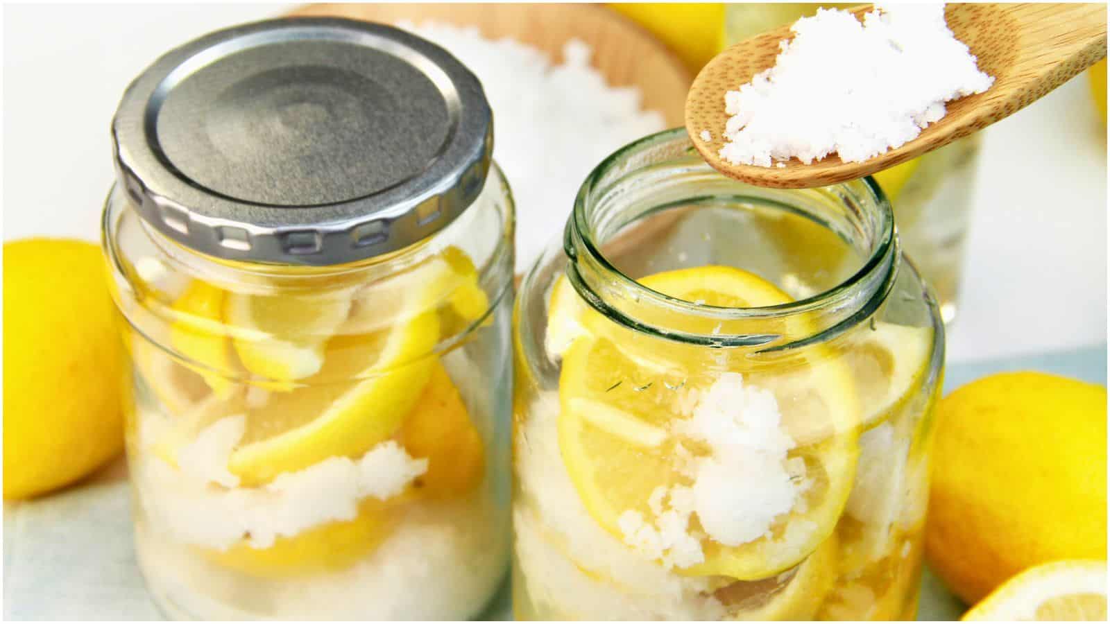 Two glass jars filled with lemon slices and a white granular substance, likely salt. The left jar is closed with a metal lid, while the right jar is open. A wooden spoon with white granules hovers above the open jar. Lemons are scattered around the jars.