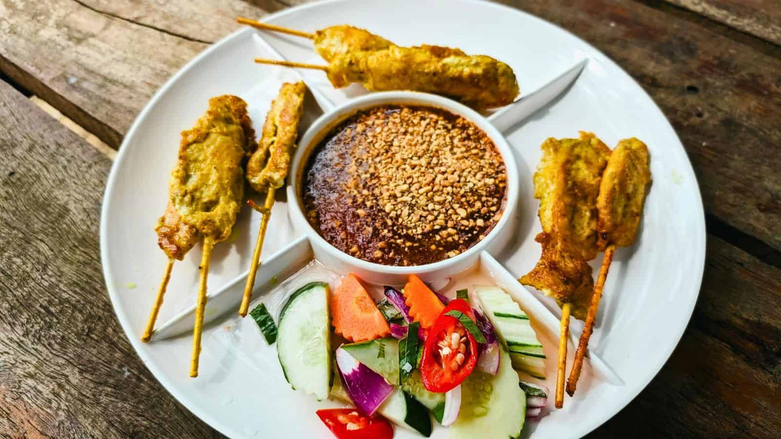 A white plate with five skewers of grilled meat arranged around a bowl of peanut sauce, sprinkled with crushed peanuts. The plate also includes a side salad with cucumber, tomato, red onion, and red chili slices on a wooden table.