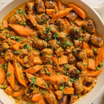 A white pan filled with a stew featuring browned meat chunks, carrot slices, and green peas in a thick, golden-brown sauce. Fresh herbs are sprinkled on top. A small bowl with chopped herbs is visible in the background—perfect for your dinner recipes collection and breaking that takeout addiction.