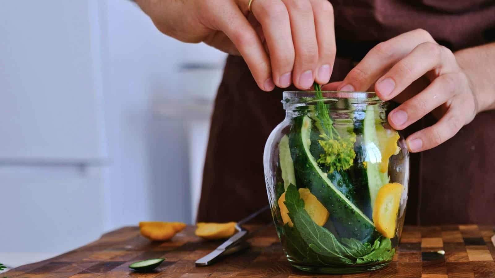 A person wearing a brown apron uses both hands to place leafy greens, cucumber, and other vegetables into a glass jar on a wooden cutting board. Sliced vegetables and a knife are also on the cutting board.