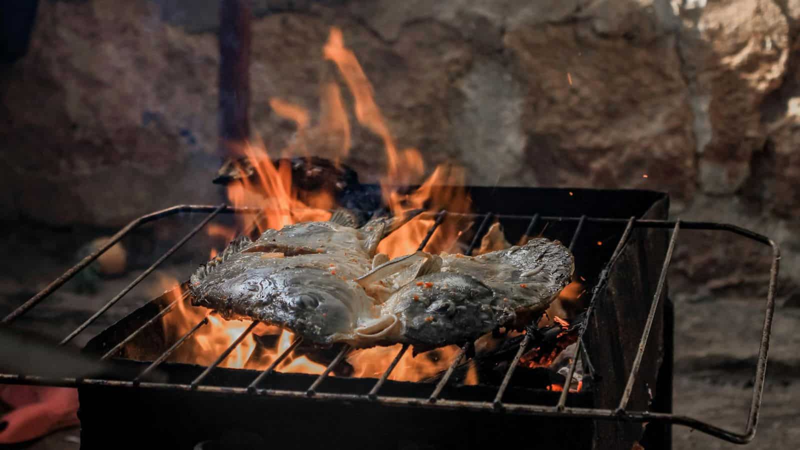 Two fish are being grilled over an open flame on a metal grill. The whole fish, positioned side by side, generate a charred and smoky effect as they cook. A stone wall is visible in the background.