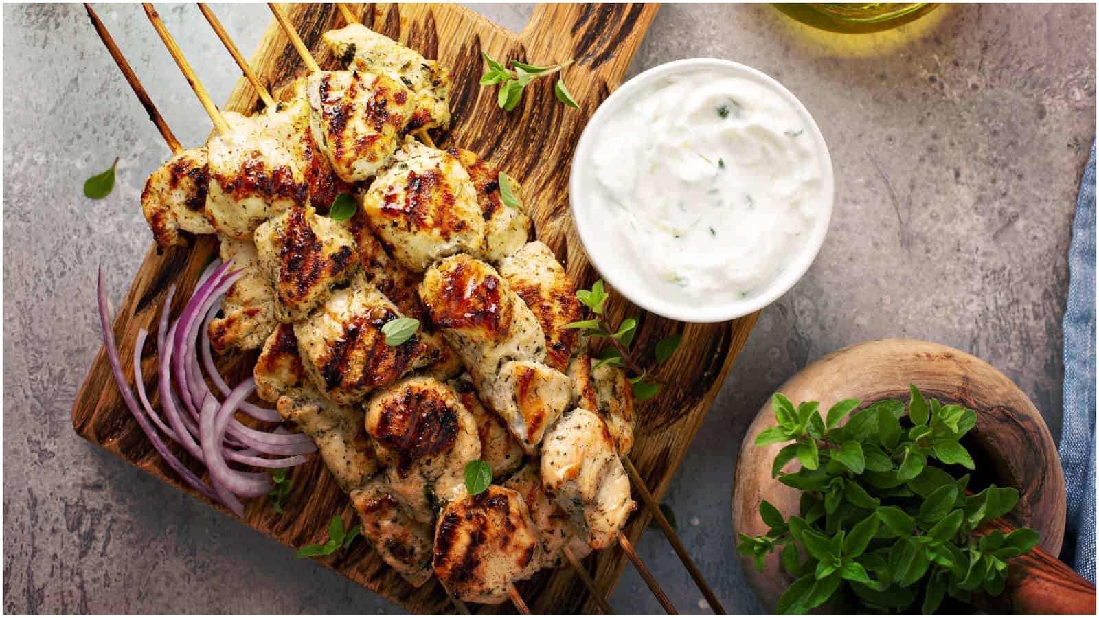 Three grilled chicken skewers on a wooden board are garnished with herbs and placed beside a small bowl of white sauce. Sliced red onions and fresh herbs are visible close by. A partial view of an olive oil bottle and a cloth napkin can be seen.