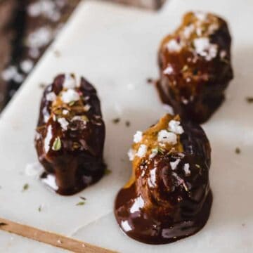 Three chocolate-covered dates topped with sea salt and herbs rest on a white marble serving board, embodying the perfect summer dessert. The background features a wooden table sprinkled with scattered sea salt, adding a rustic charm to this delightful treat.