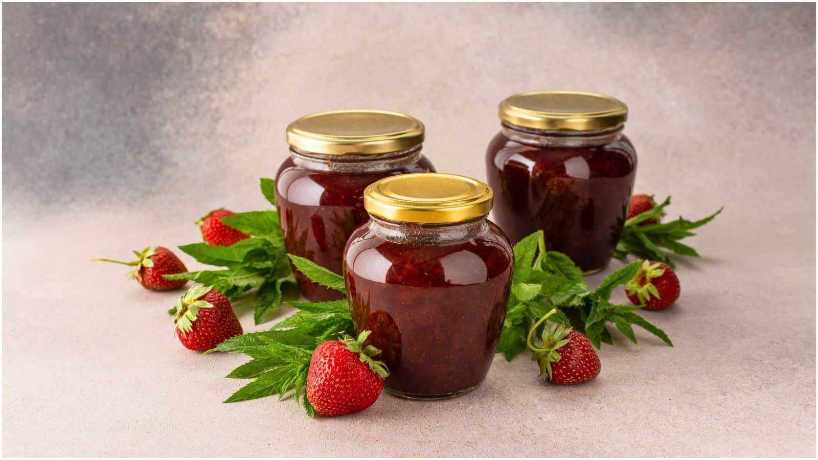 Three glass jars filled with strawberry jam and sealed with golden lids are surrounded by fresh strawberries and green leaves all arranged on a neutral-colored surface. The background is blurred with shades of gray and brown.