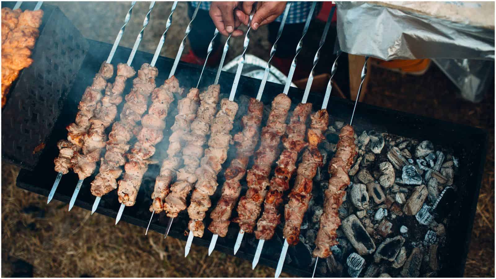 Close-up of several skewers of meat being grilled over hot coals. A person is seen turning one of the skewers. The marinated meat appears slightly charred from grilling. Smoke is visible rising from the grill.