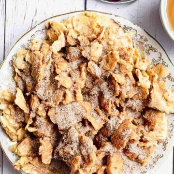 A plate of Kaiserschmarrn, an Austrian shredded pancake, is served with powdered sugar. A cup of tea and a small bowl of sauce are placed next to the plate on a white wooden surface. A fork and knife are positioned to the left, resting on a grey napkin.