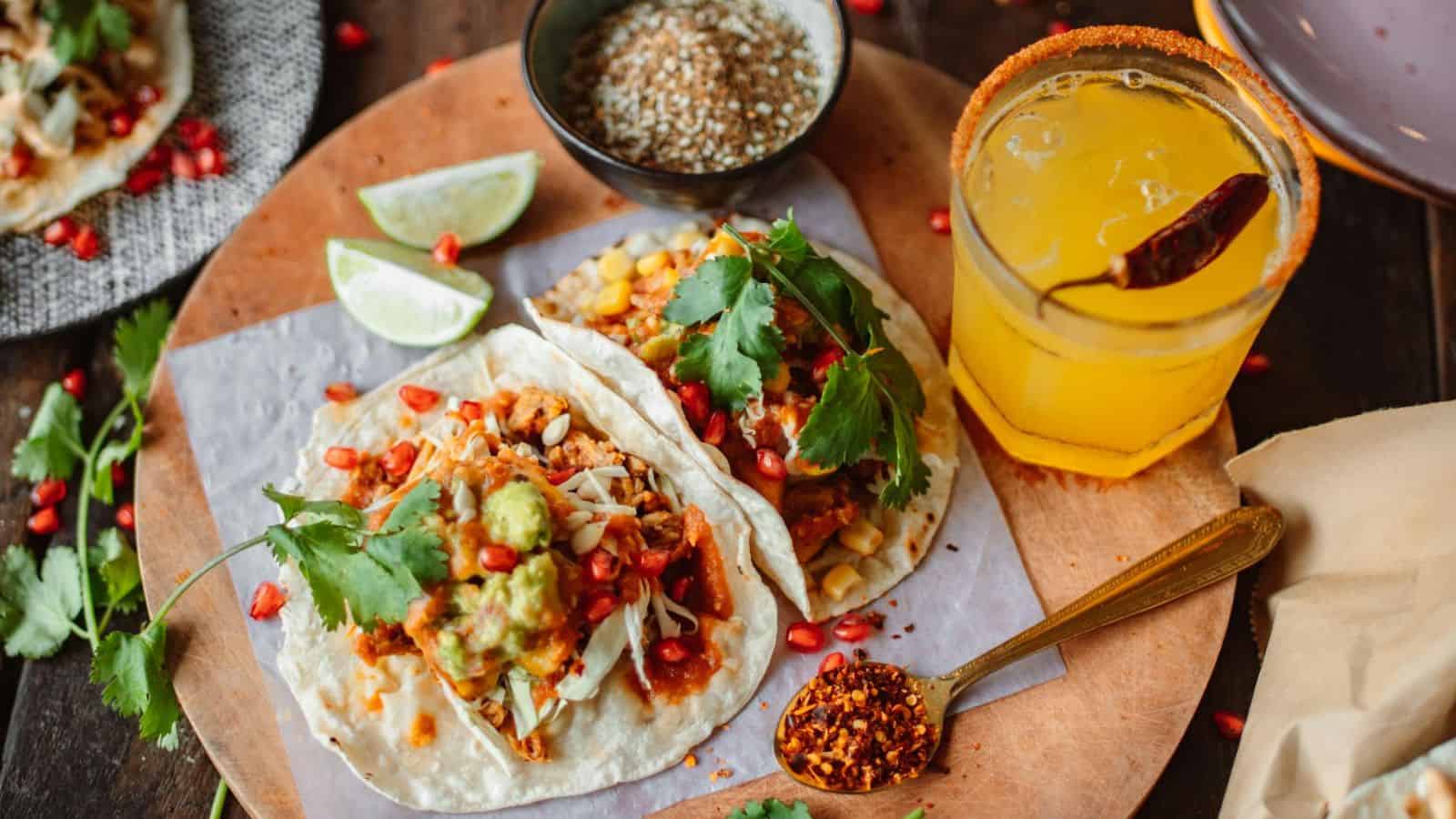 A wooden board holds two tacos filled with ingredients like guacamole, pomegranate seeds, and garnished with cilantro, accompanied by lime wedges, a bowl of sauce, and a drink with a chili garnish. Inspired by iconic street foods from around the world, there's also a spoon with red spice next to the tacos.