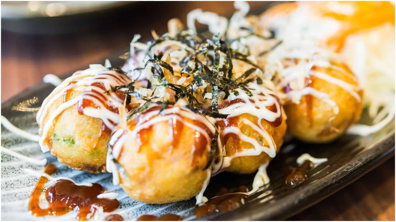 A close-up of a dish featuring four takoyaki balls. They are drizzled with mayonnaise, a savory brown sauce, and topped with thin strips of seaweed and bonito flakes. The takoyaki balls are arranged on a dark, rectangular plate.
