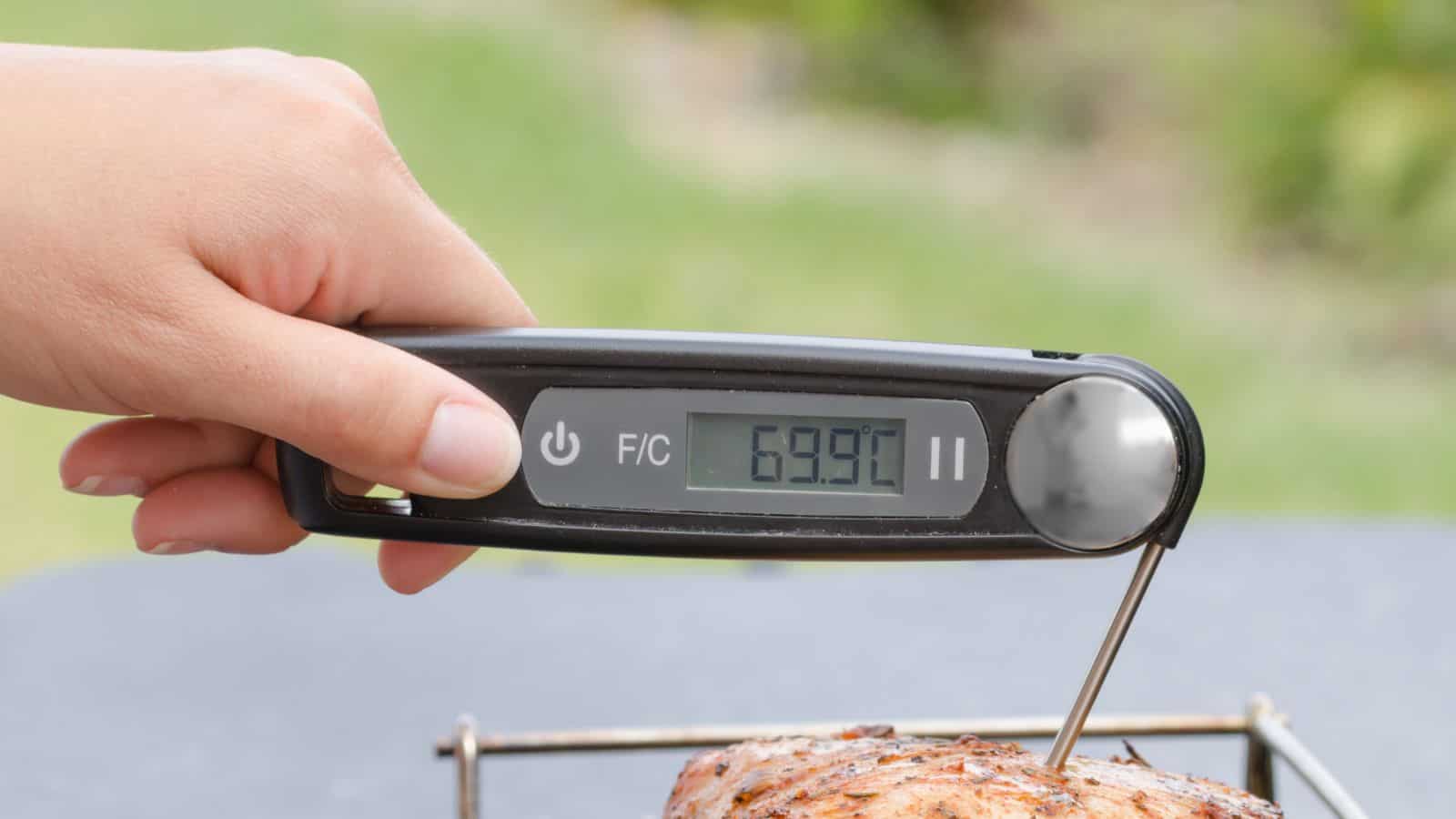 A hand is holding a digital meat thermometer, an essential tool for any grill master, displaying a temperature of 69.9°C. The thermometer is inserted into a piece of meat resting on the grill.