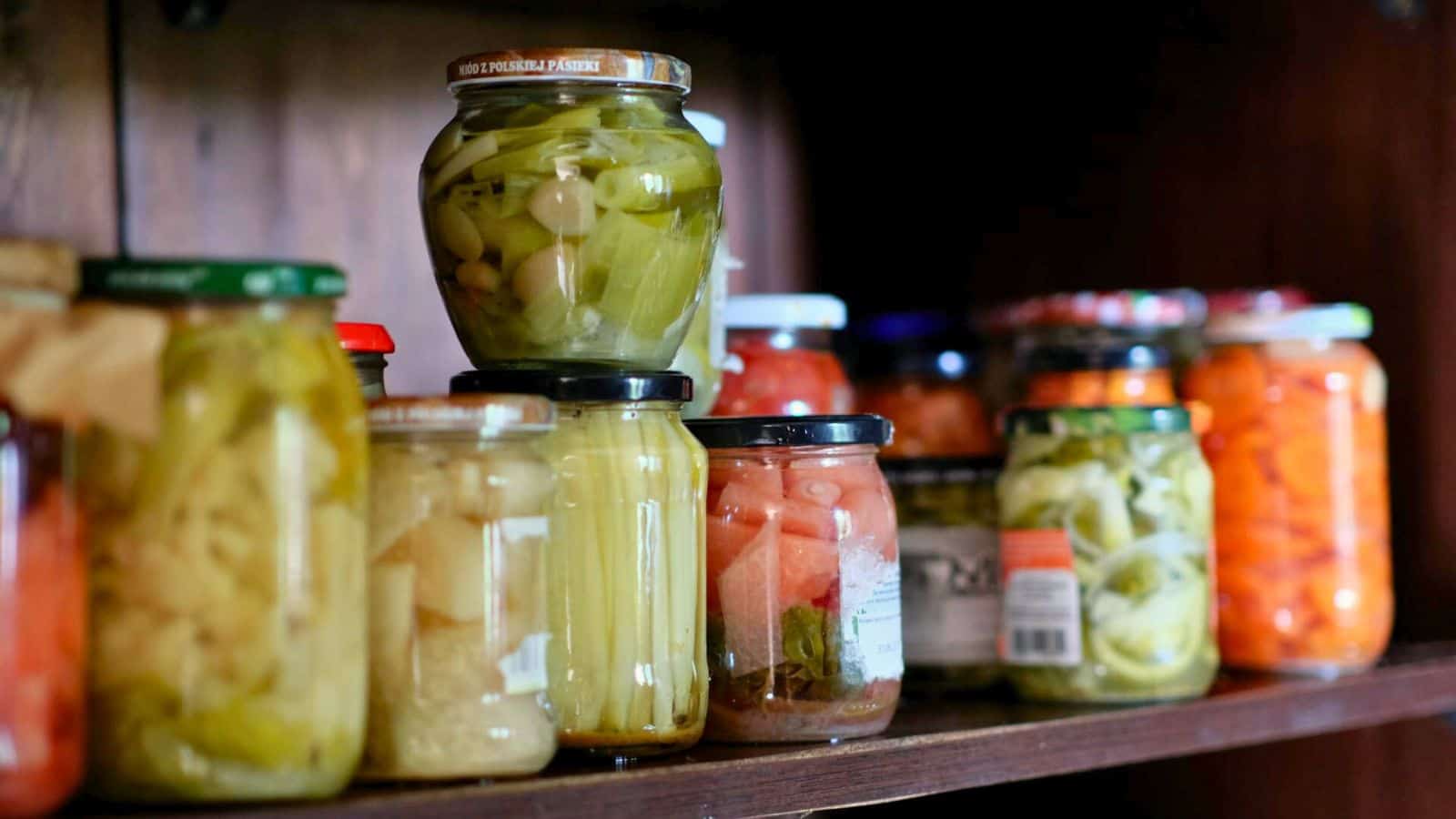 A variety of glass jars filled with pickled vegetables sit on a wooden shelf. The jars contain different colors and types of vegetables, including green, yellow, orange, and red varieties, each sealed with a metal lid. The shelf is part of a larger wooden structure.