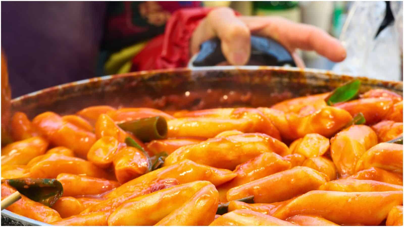 A close-up of a large pan filled with tteokbokki, one of the iconic street foods from Korea, consisting of stir-fried rice cakes in a spicy red sauce. The dish includes green vegetables and is being stirred by a person holding a utensil.