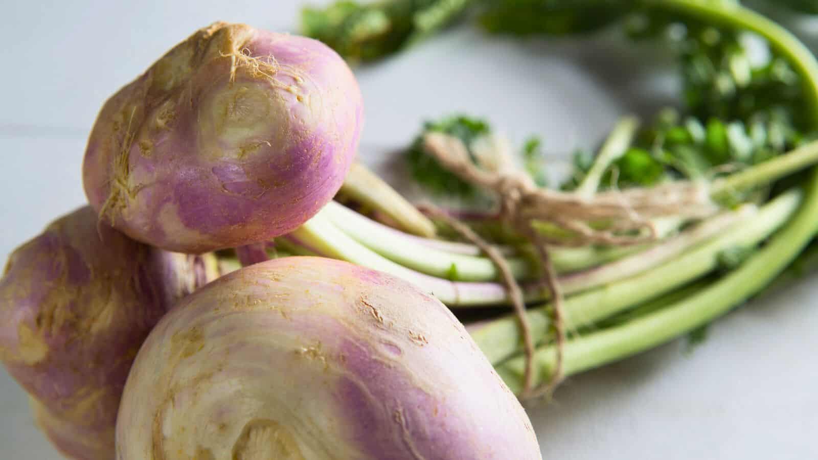Three rutabagas with purple tops and green leafy stems sit on a white surface. The stems, tied together with a piece of twine, hint at their freshly harvested state. The roots have uneven surfaces and some dirt remains on them.