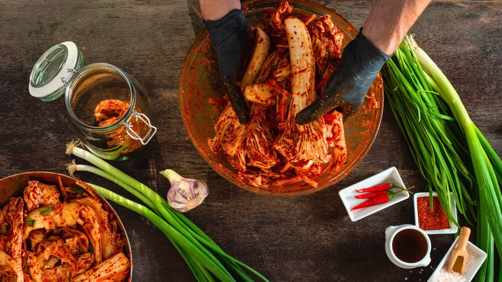 Hands wearing black gloves are mixing kimchi in a large bowl on a wooden surface. Green onions, garlic, a jar of kimchi, red chili peppers, a small bowl of sauce, red chili powder, and salt are arranged around the workspace.