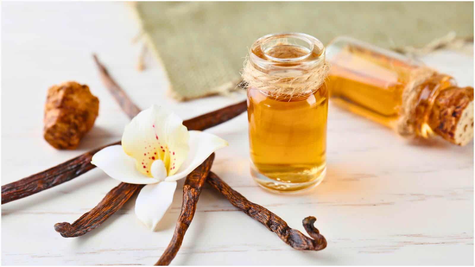A small glass vial filled with a light brown liquid is wrapped with twine. Nearby are three vanilla beans, a white orchid flower, and a piece of burlap on a light wooden surface. A second vial lies in the background, creating an artisanal display.