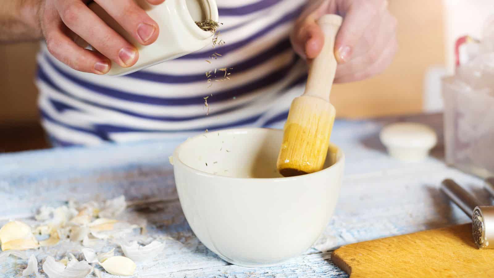 A person in a striped shirt sprinkles herbs from a small container into a white bowl while holding a wooden pestle. Garlic cloves and their peels are scattered on the surface nearby.