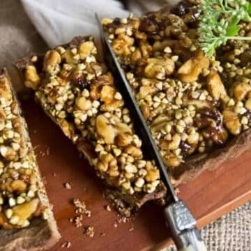 A rectangular tart with a nut topping is being sliced with a knife. The tart features a mixture of different nuts and possibly a drizzle of syrup. It is presented on a wooden board with a garnish, and two slices are partially separated from the main tart.