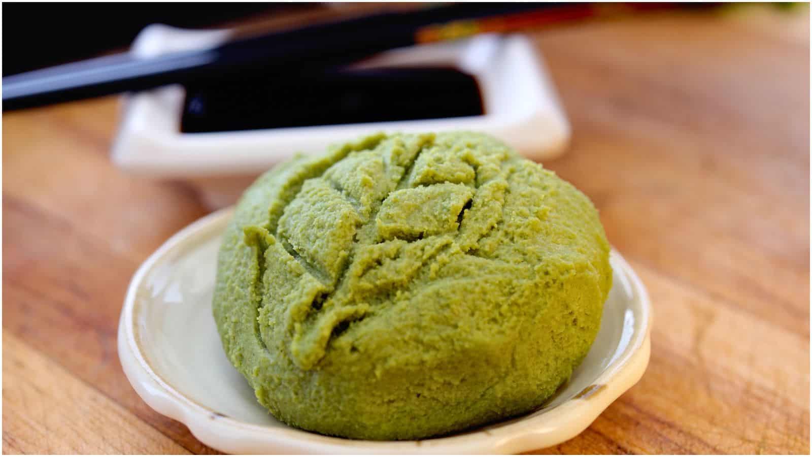 A close-up of a dish containing wasabi, placed on a small white plate. In the background, there are a pair of black chopsticks and a white dish with soy sauce, resting on a wooden surface.