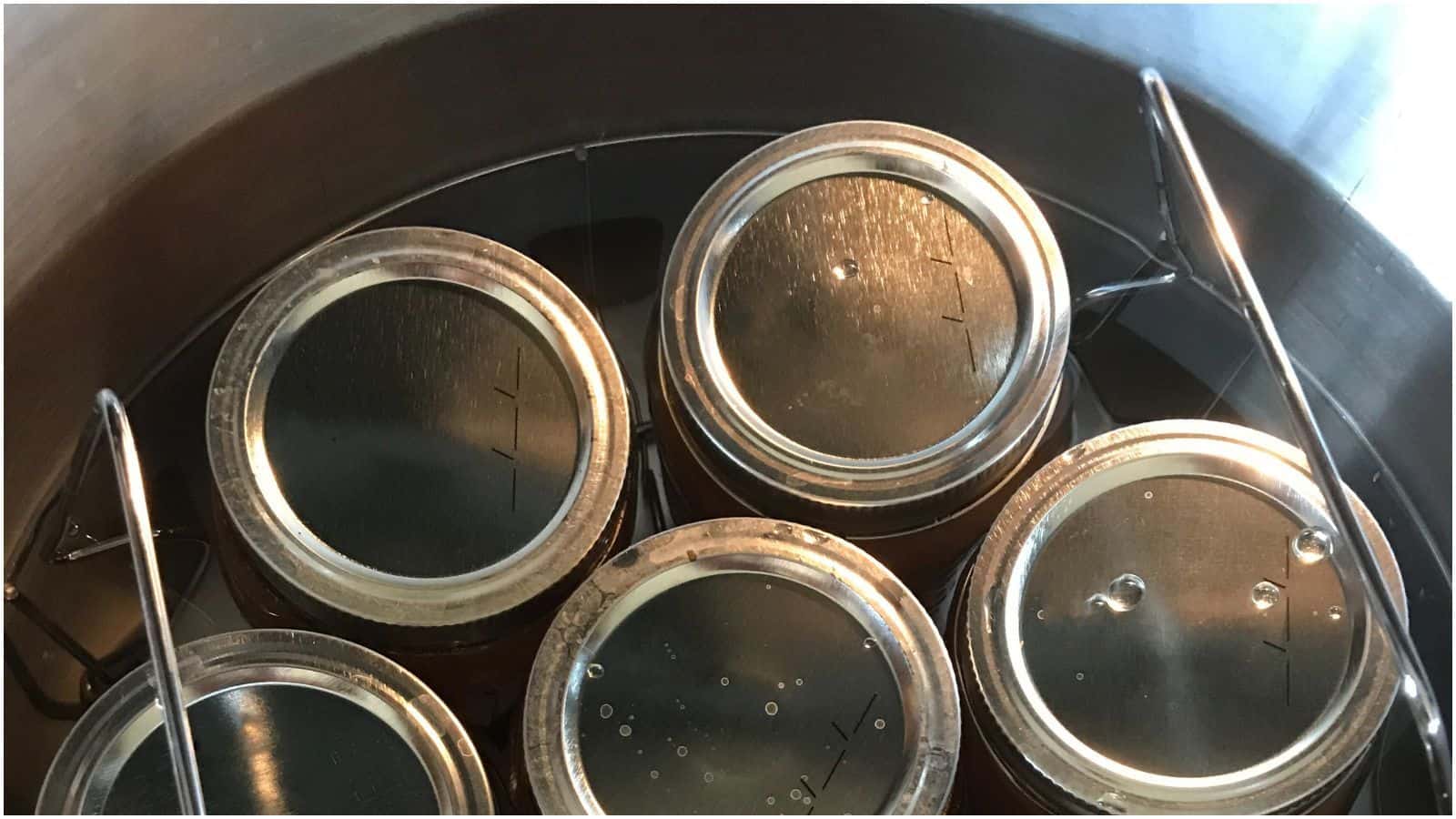 Five sealed mason jars are partially submerged in a pot of water and positioned in a circular manner. The jars are supported by a metal rack inside the pot.