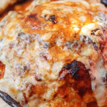 A baked cauliflower head is covered with melted cheese and browned edges, presented in a glass baking dish. The dish has a reddish sauce that appears to be tomato-based, surrounding the base of the cauliflower.