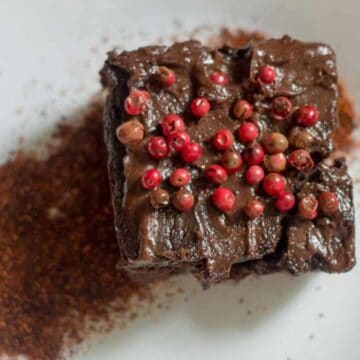 A chocolate cake square garnished with pink and red peppercorns sits on a white plate. Beside the cake is a small heap of ground spices or cocoa powder. The background shows a wooden table surface.