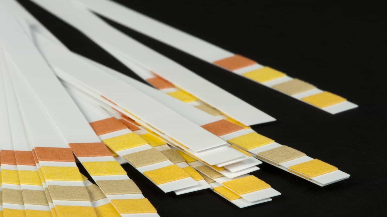 A close-up image of several white pH test strips on a black surface. The strips have colored indicator sections in shades of yellow and orange, used to test the acidity or alkalinity of substances.