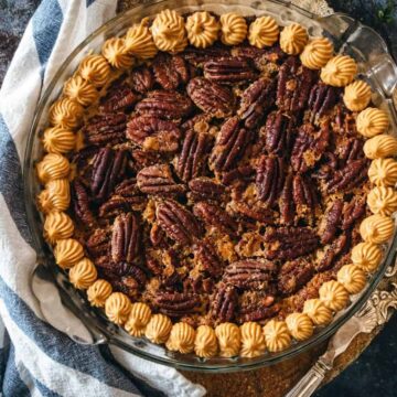 A pecan pie in a glass dish with decorative piped cream along the edge, this delightful summer treat is set on a dark, textured surface next to a striped cloth napkin and a vintage metal pie server.