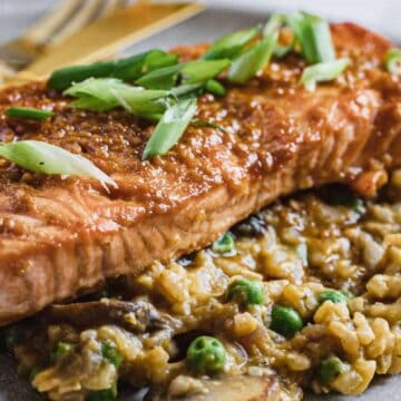 A plate of food featuring a piece of seared salmon garnished with sliced green onions, served over a bed of risotto with peas and mushrooms. A fork and knife are placed beside the dish.