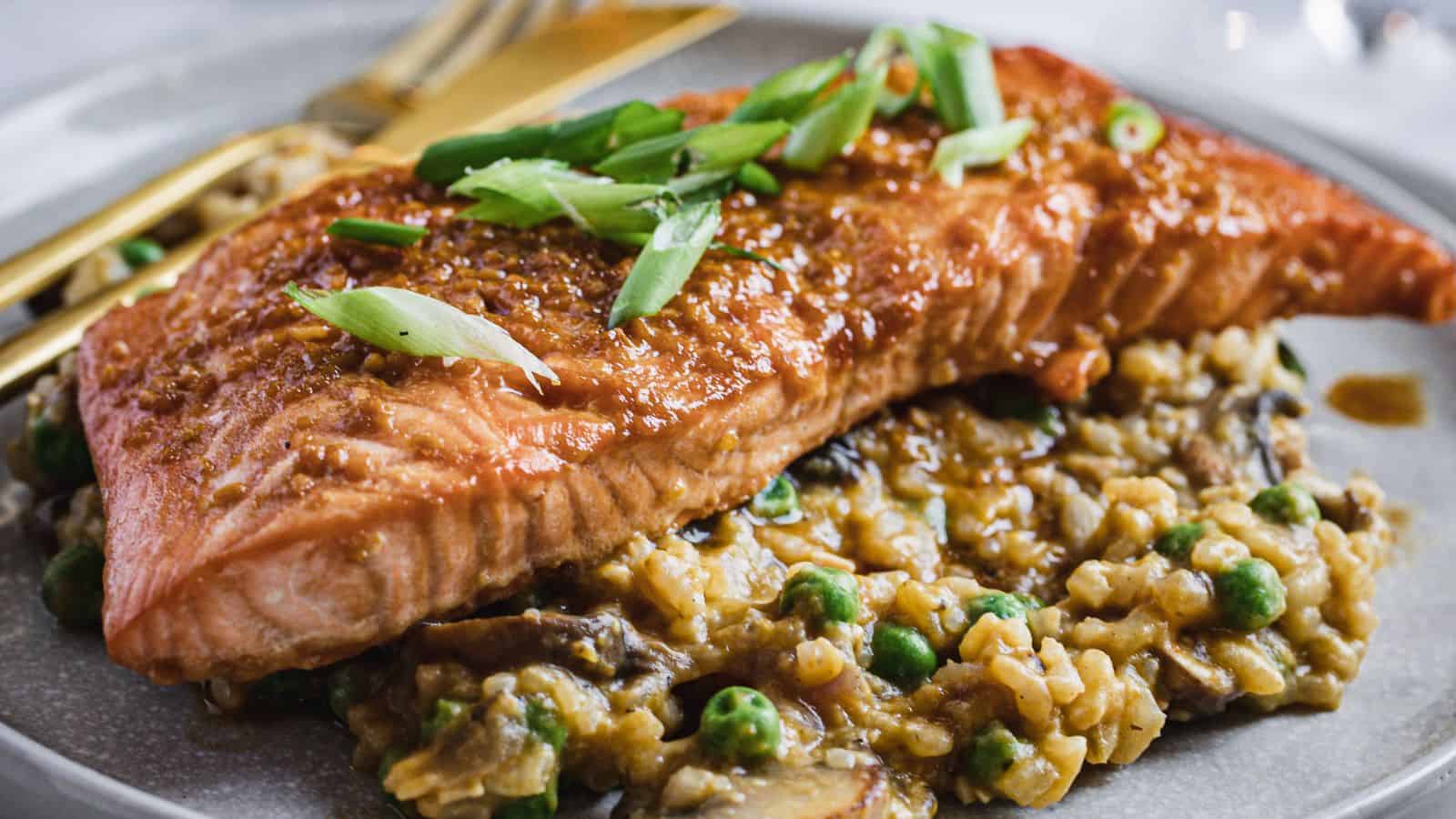 A teriyaki salmon fillet topped with sliced green onions is placed on a plate, resting on a bed of creamy risotto mixed with peas and mushrooms. A gold fork and knife are positioned beside the salmon.