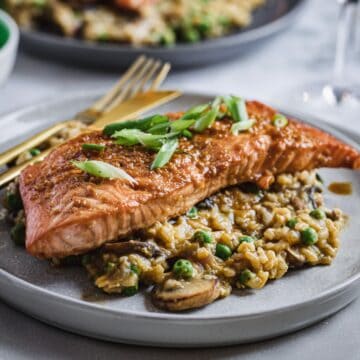 A plated meal features a seared teriyaki glazed salmon fillet topped with green onions, resting on a bed of mushroom risotto with peas. Behind, a similar dish is visible out of focus. A fork and knife are placed beside the plate, and a glass of white wine is in the background.