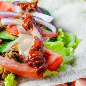 A close-up of a wrap filled with shredded spiced meat, sliced tomatoes, cucumbers, red onions, and lettuce drizzled with a red sauce and white dressing. The wrap appears to be made of a thin, white flatbread.