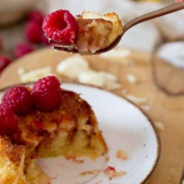 A small plate holds a partially eaten piece of cake topped with fresh raspberries. A hand is holding a fork with a bite of the cake and a raspberry. Several raspberries are scattered on a wooden surface in the background.