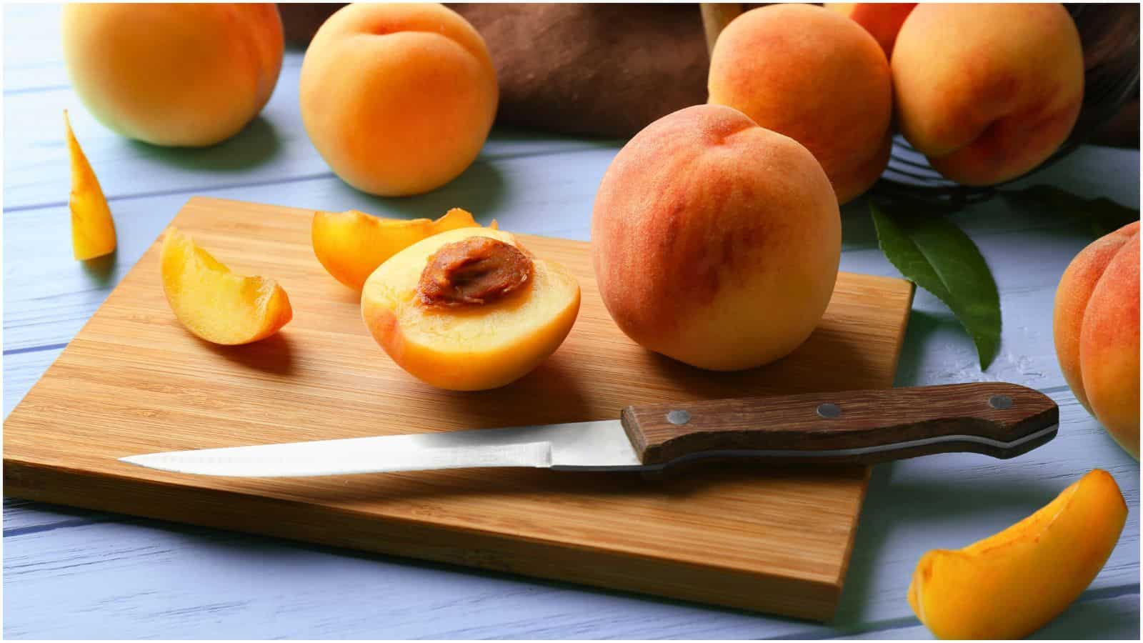 A wooden cutting board with a serrated knife, several whole peaches, and some peach slices. One peach is halved, revealing the pit. The background is a light blue surface scattered with more whole peaches and a brown cloth—a perfect setup for learning how to freeze peaches effectively.