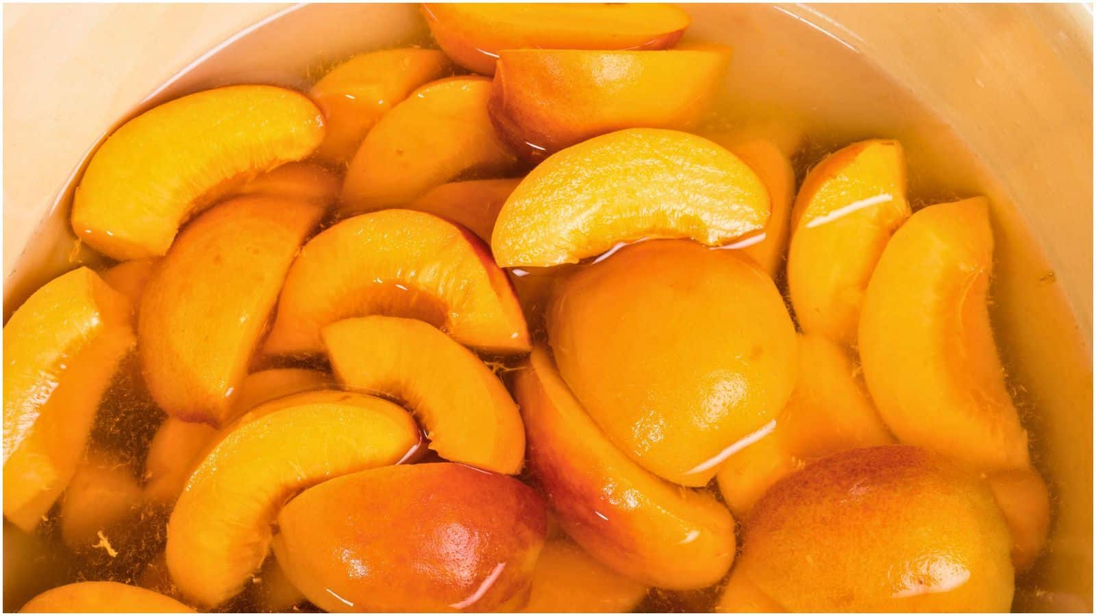 Close-up of sliced peaches submerged in a liquid, possibly syrup or juice, within a light-colored container. The peaches are a vibrant orange and appear to be preserved or canned. Understanding how to freeze peaches can help maintain their fresh color and flavor.