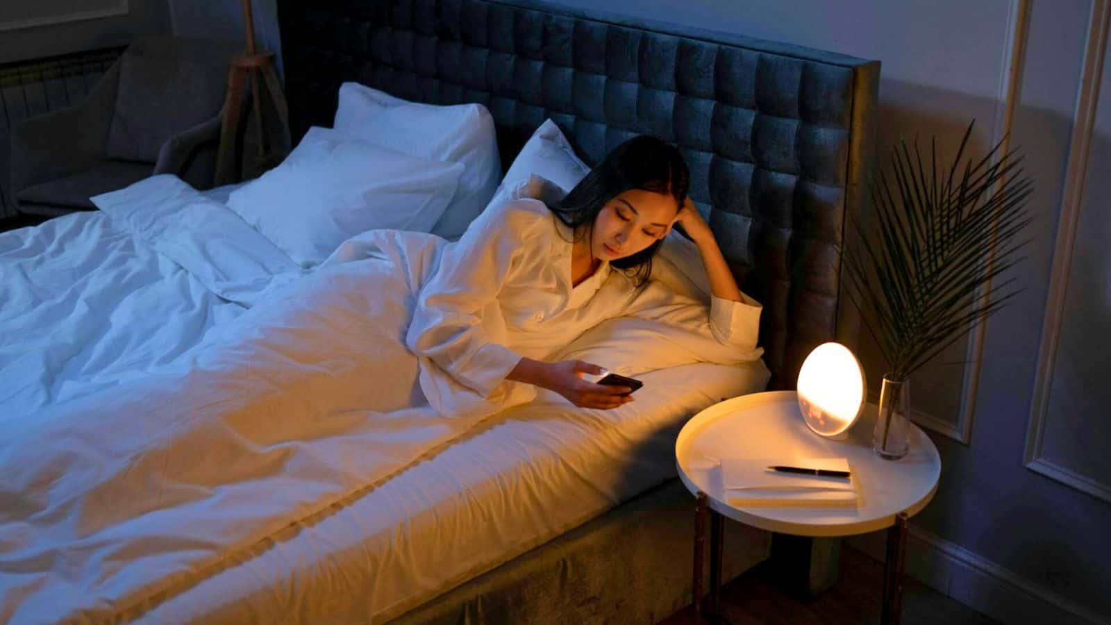 A woman in a white shirt lies on a bed using a smartphone. The room is dimly lit, with a round nightstand beside the bed holding a modern lamp, a book, and a pen. A plant is visible on the nightstand. The bed has white linens and a padded headboard.