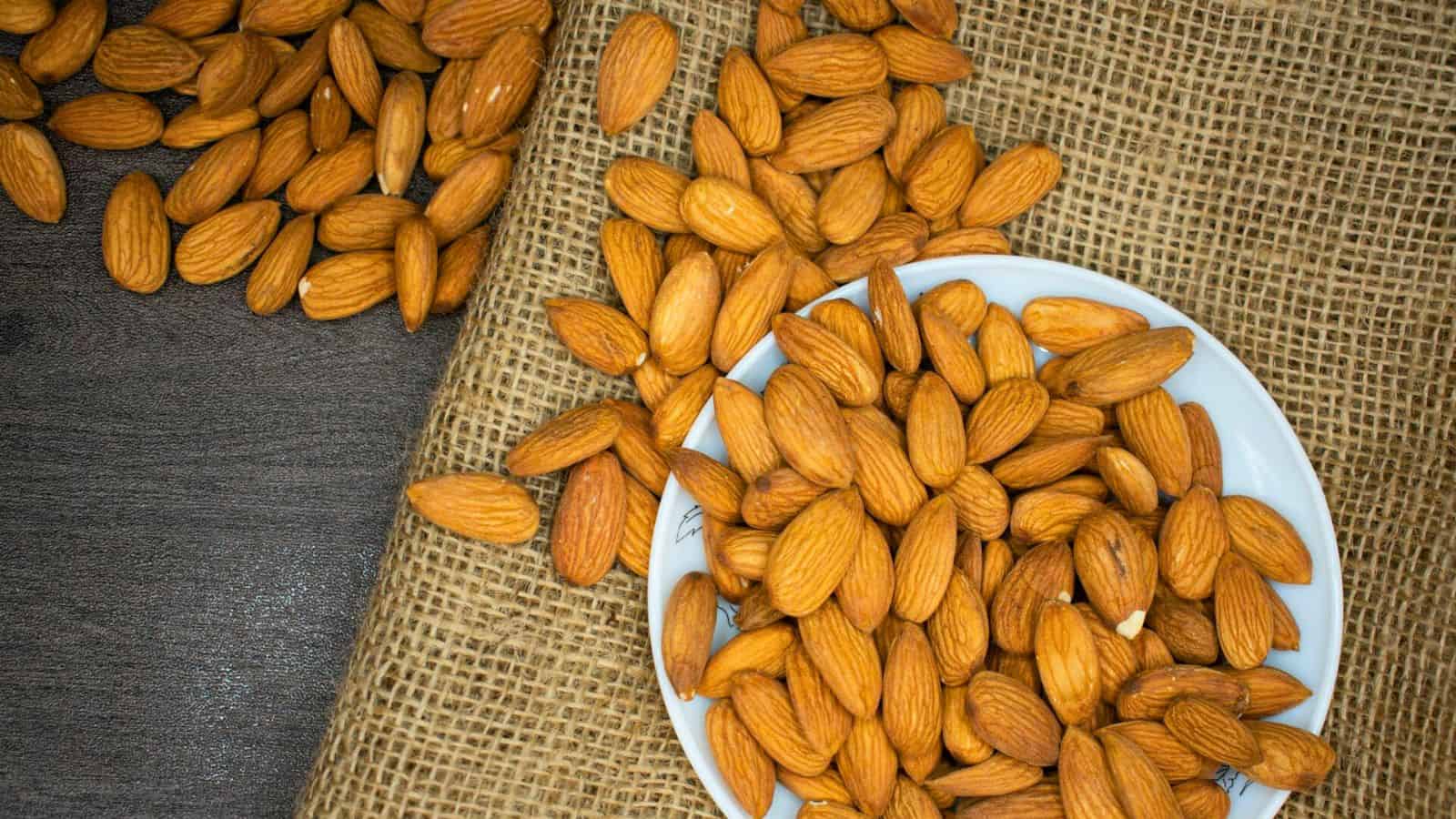 A bowl filled with almonds is placed on a piece of burlap fabric. More almonds are scattered around the bowl and on the burlap. There is a dark wooden surface underneath the burlap. The almonds appear dry and have a natural brown color.